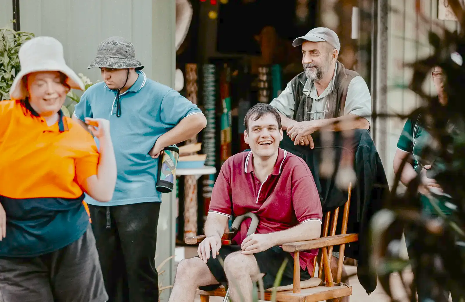 A group of adults, some wearing hats, gather outdoors. One man sits and smiles in the centre as others stand nearby, chatting and laughing—a warm moment at a Compass Disability Services event. Shelves and a partially open building appear in the background.