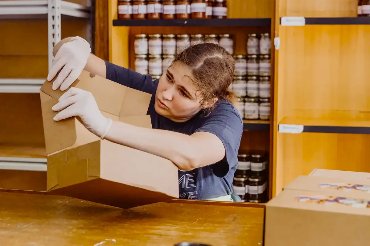A person wearing white gloves is assembling a cardboard box at a wooden table in Harvest Kitchen, with shelves of jars visible in the background.