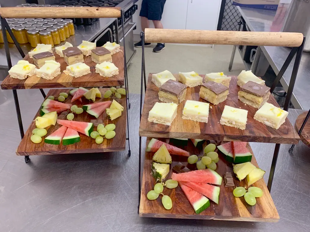 Two wooden serving trays with handles display assorted dessert bars on the top shelves and Harvest Kitchen’s fresh fruit slices, including watermelon, pineapple, and green grapes, on the bottom shelves in a commercial kitchen setting.