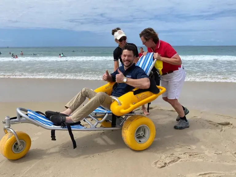 A man sits smiling and giving a thumbs up in a beach wheelchair with yellow wheels, while two people from Disability Services stand behind him on the sand, helping him near the sea on a partly cloudy day.