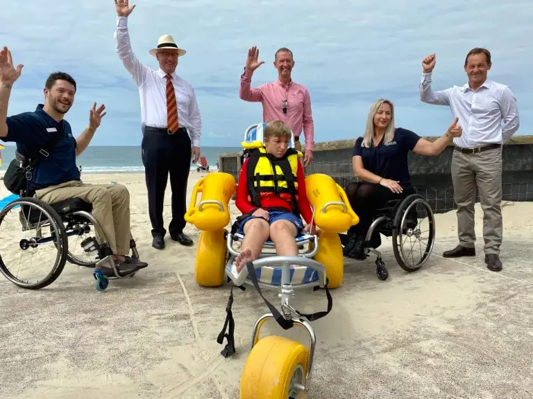 A group of five adults smiles and waves by the beach; two use wheelchairs, and a boy in a yellow life jacket sits in a Compass beach wheelchair with large yellow wheels.