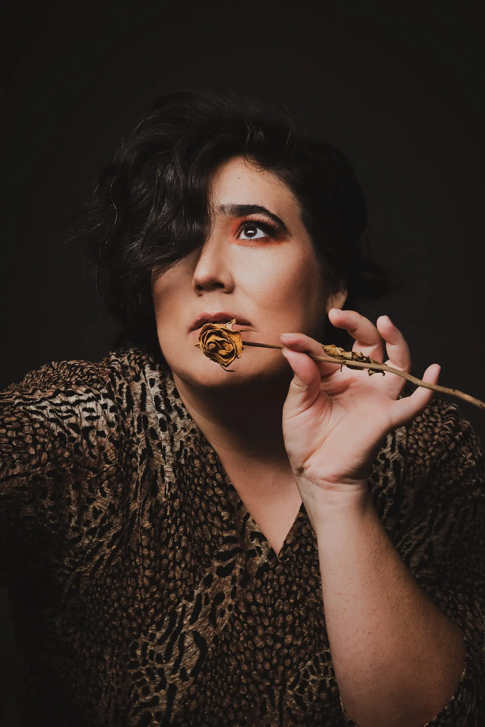 A person with short, dark wavy hair and dramatic make-up gazes upward as if pondering "What If," holding a dried, wilted rose near their mouth. They wear a brown leopard-print top against a dark background.