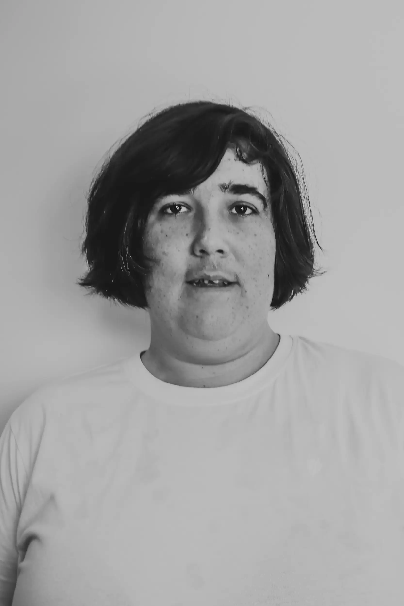 A person with short dark hair and freckles is wearing a plain white T-shirt, standing against a light-coloured background. The black and white image evokes a sense of contemplation, as if pondering a "What If" moment in time.
