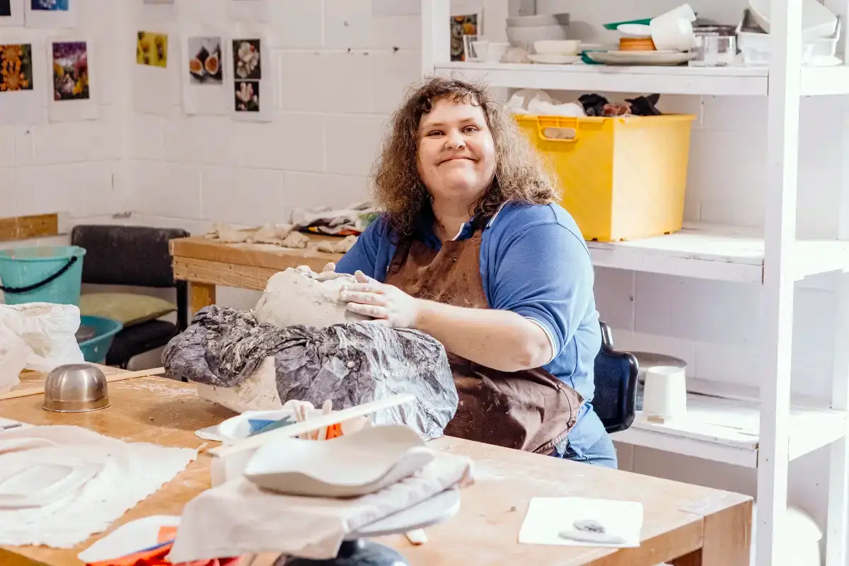 A smiling person wearing a blue shirt and brown apron is sculpting a clay head in an art studio filled with pottery tools and finished ceramics, inspired by the spirit of What If creativity.