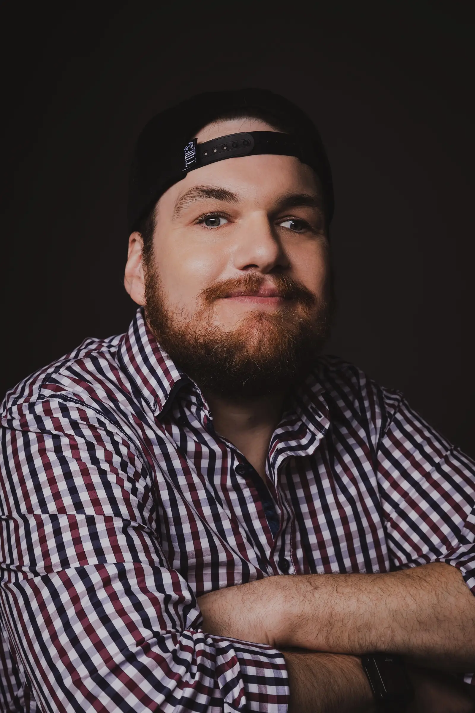 A bearded man wearing a checked shirt and a backwards black cap smiles slightly while sitting with his arms crossed against a dark background, as if pondering "What If" moments or contemplating new directions like a compass guiding Disability Services.
