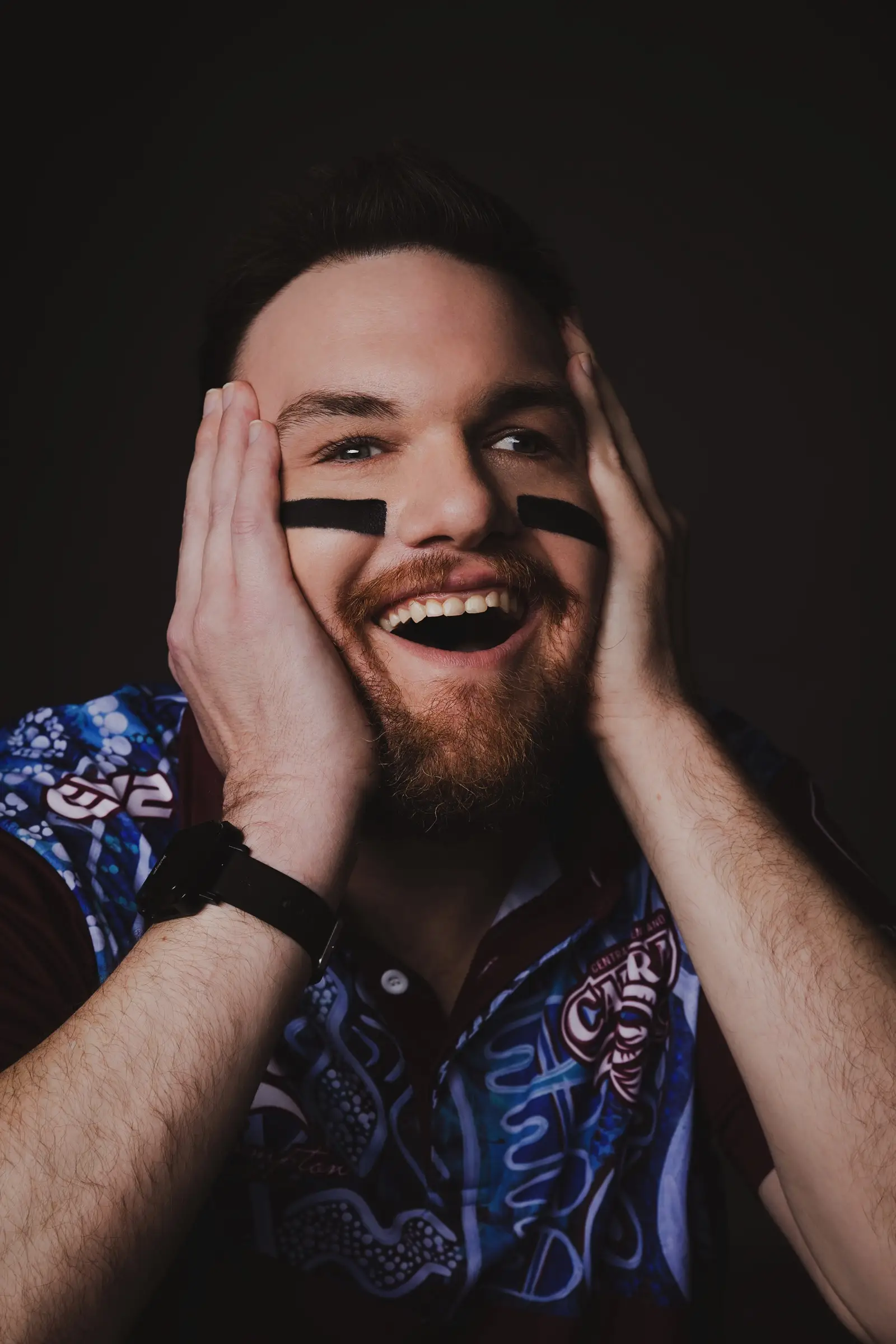 A bearded man with black face paint under his eyes smiles widely, holding his face with both hands. He wears a patterned blue and purple shirt and a black Compass watch, set against a dark background.