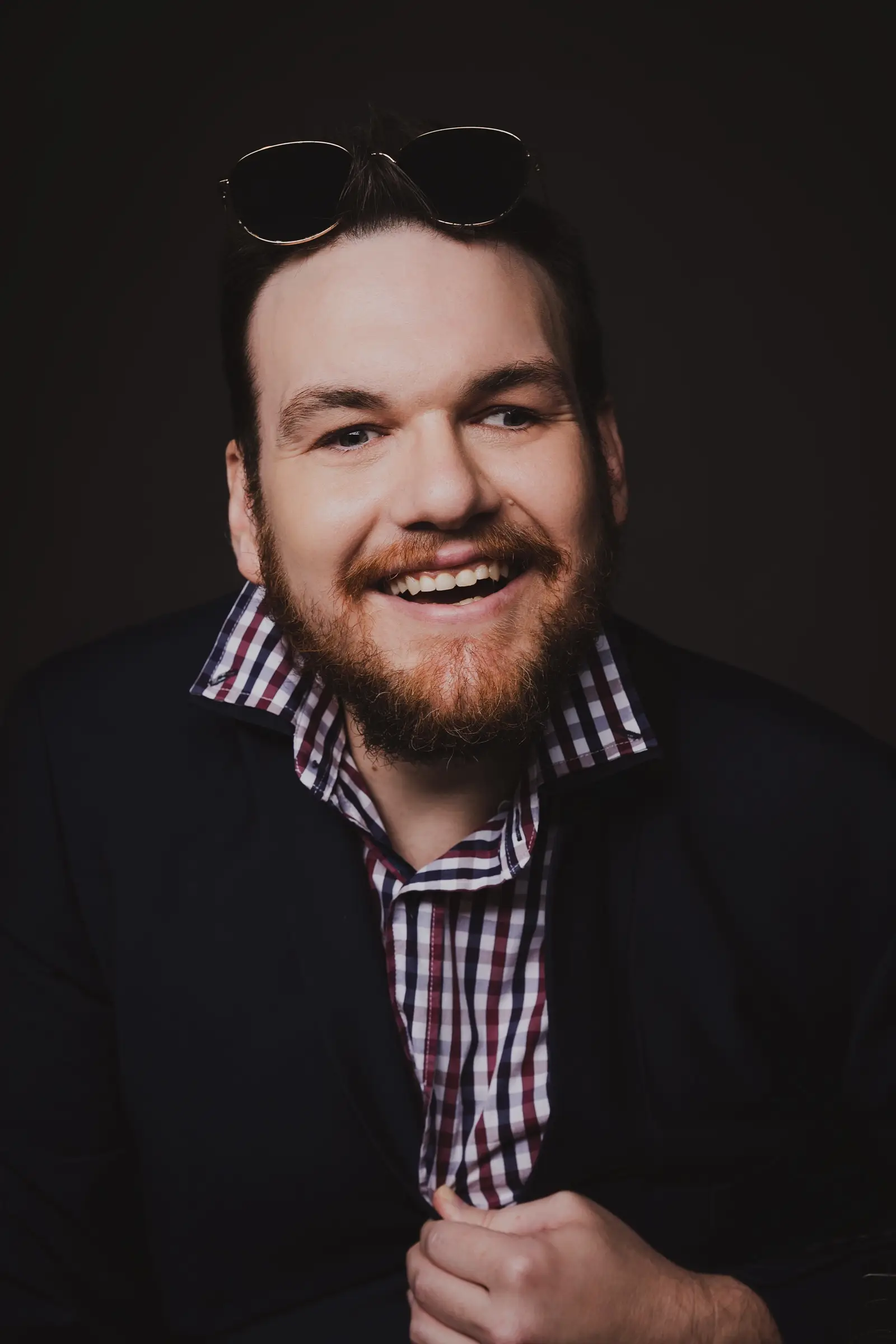 A smiling man with a beard wears a chequered shirt under a dark jacket. With sunglasses resting on his head, he poses confidently against a dark background—ready to guide others like a compass in Disability Services.
