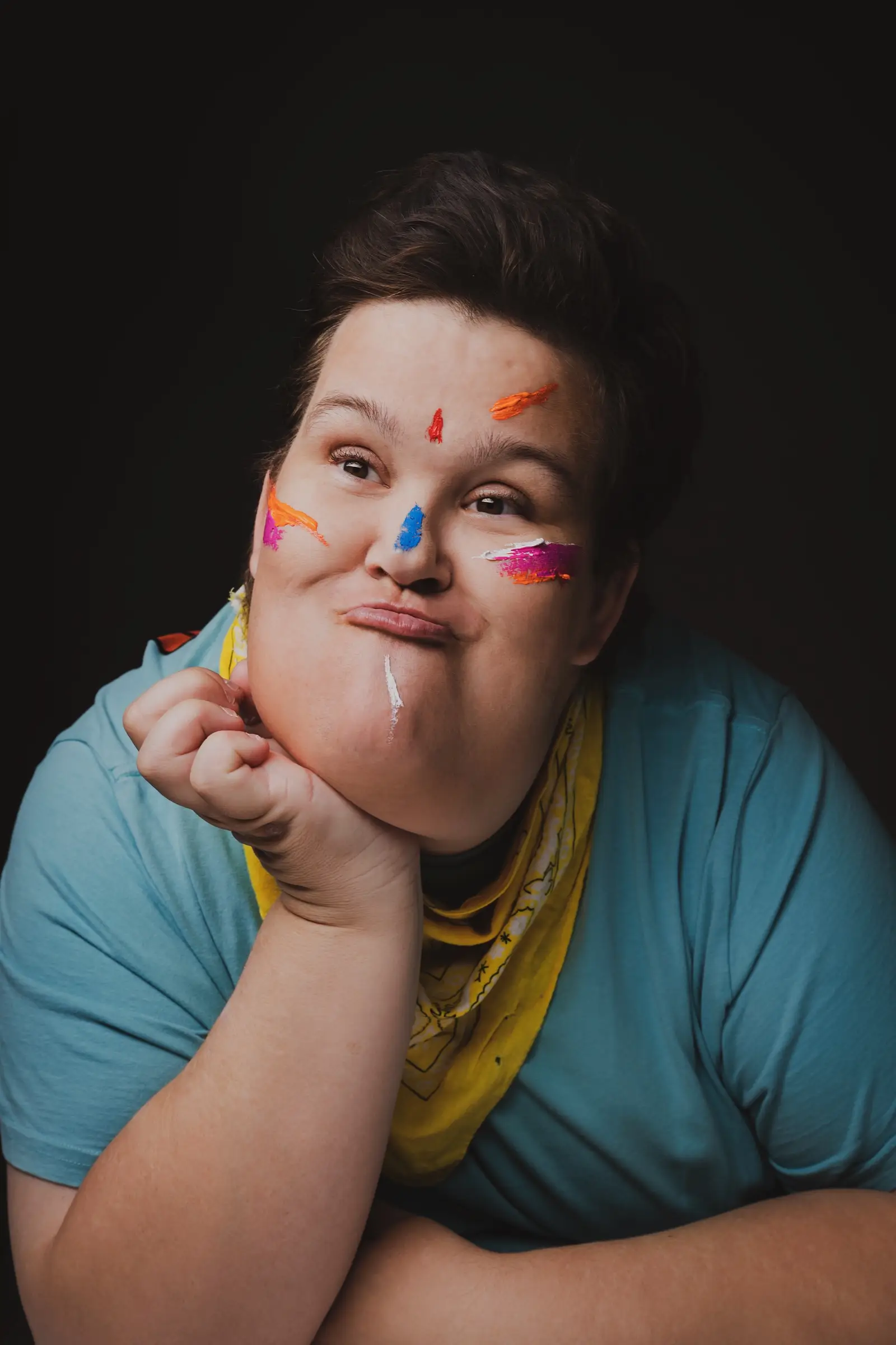 A person with short dark hair and a blue shirt, wearing a yellow bandana, rests their chin on their hand and looks upward—as if contemplating "What If." Their cheeks are decorated with colourful paint streaks. The background is dark.