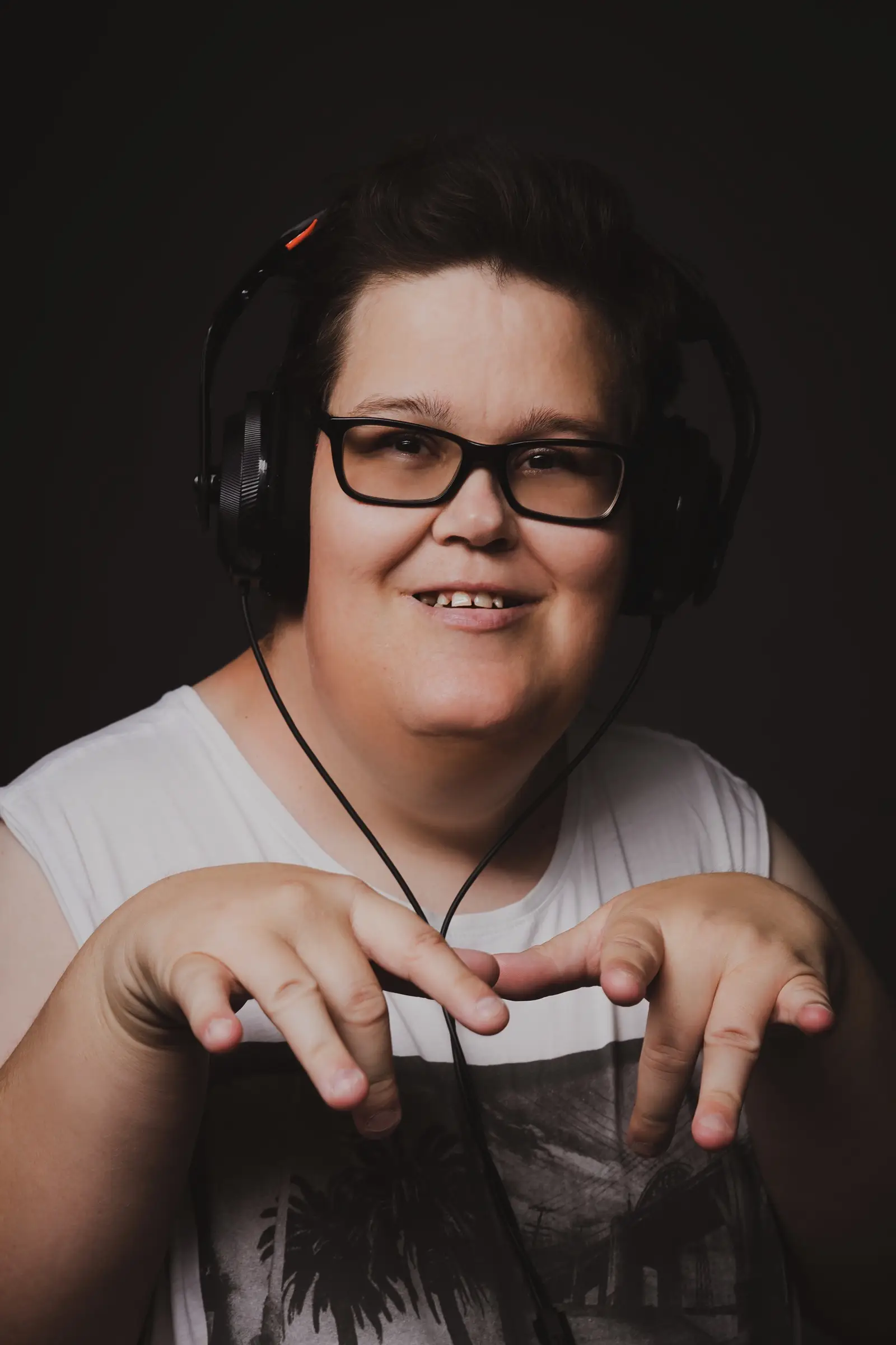 A smiling person with short dark hair, glasses, and headphones wears a white sleeveless top. They hold their hands up in front of them against a dark background, embodying the spirit of "What If" and hope inspired by Disability Services.