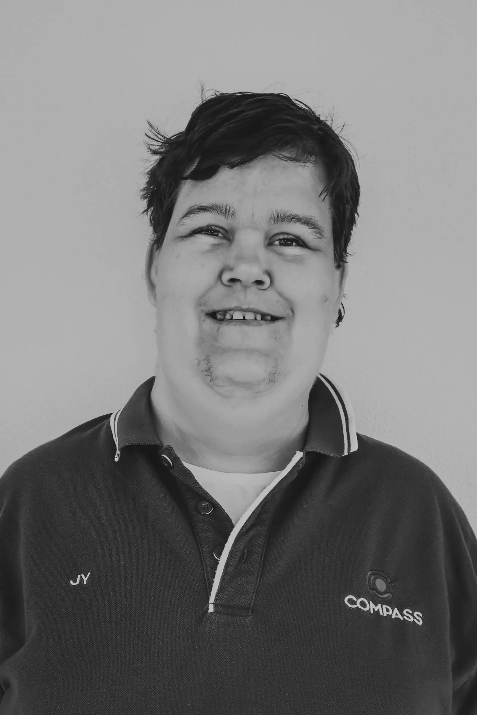A person with short dark hair, wearing a polo shirt with the words JY and Compass embroidered on it, stands against a plain background and smiles at the camera. The image, representing Disability Services, is in black and white.