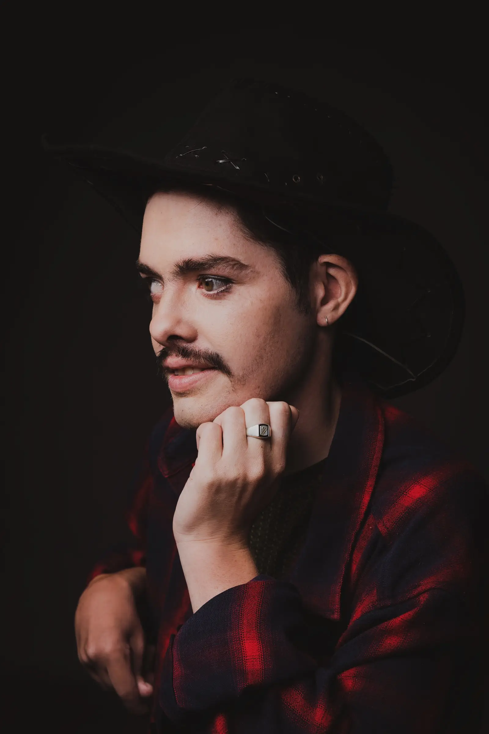 A person with light skin, a moustache, and short dark hair wears a black cowboy hat and red checked jacket. Resting their chin on one hand, they gaze thoughtfully to the side, as if pondering a "What If" scenario against a dark background.