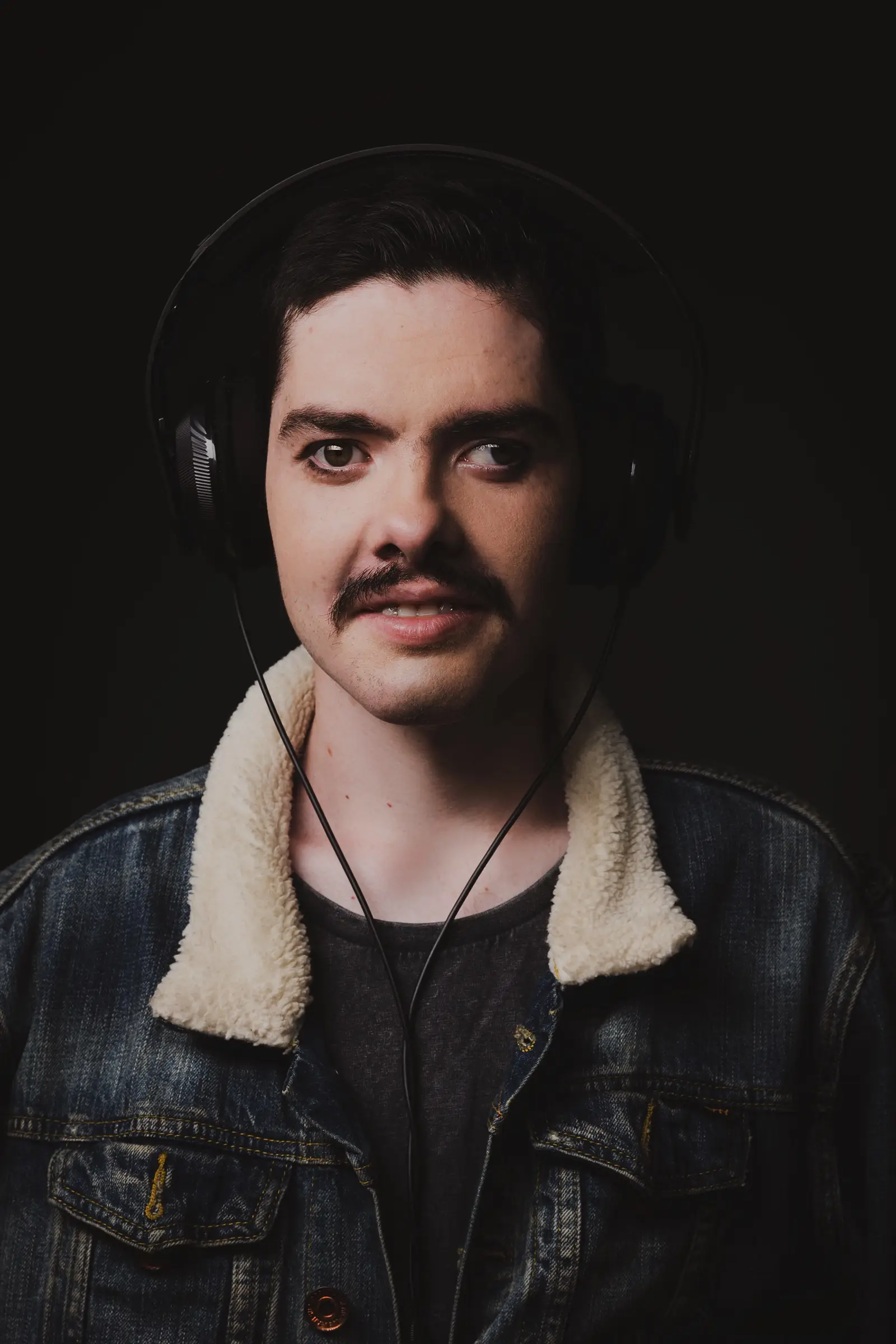 A person with short dark hair, a moustache, and headphones is wearing a denim jacket with a sheepskin collar, standing against a dark background—looking as if they're pondering "What If" moments.