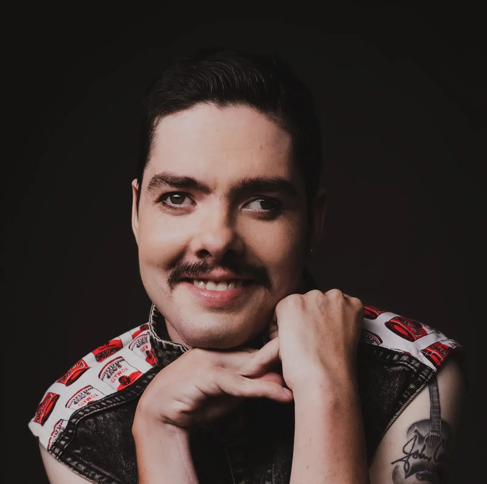 A person with short dark hair, a moustache, and light skin smiles whilst resting their chin on their hands. Wearing a Compass-themed sleeveless top with red and white patterns, they display a tattoo on their upper arm against a dark background.
