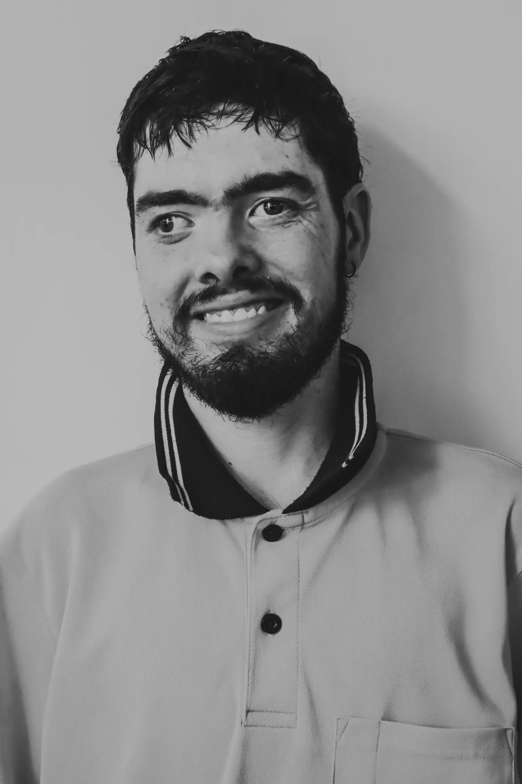 A man with short dark hair and a beard is smiling, wearing a collared shirt with a striped trim. The black and white photo features him facing slightly left against a plain background, embodying the positive spirit of Disability Services.