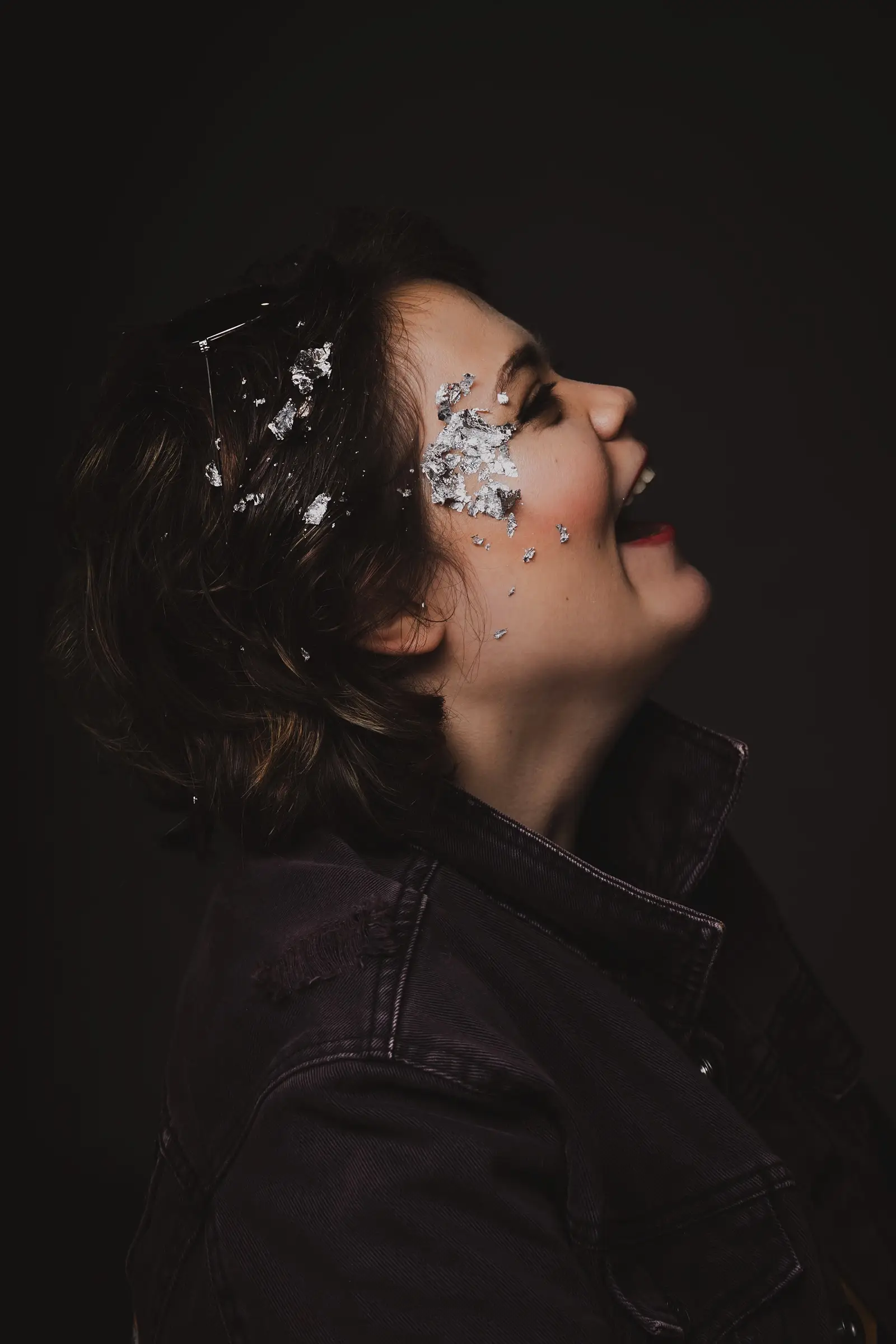 A person with short brown hair, wearing a dark jacket, smiles whilst facing sideways. Shiny silver foil pieces are artistically applied to their face and hair against a dark background, reflecting a "What If" sense of creativity and direction, like a compass.