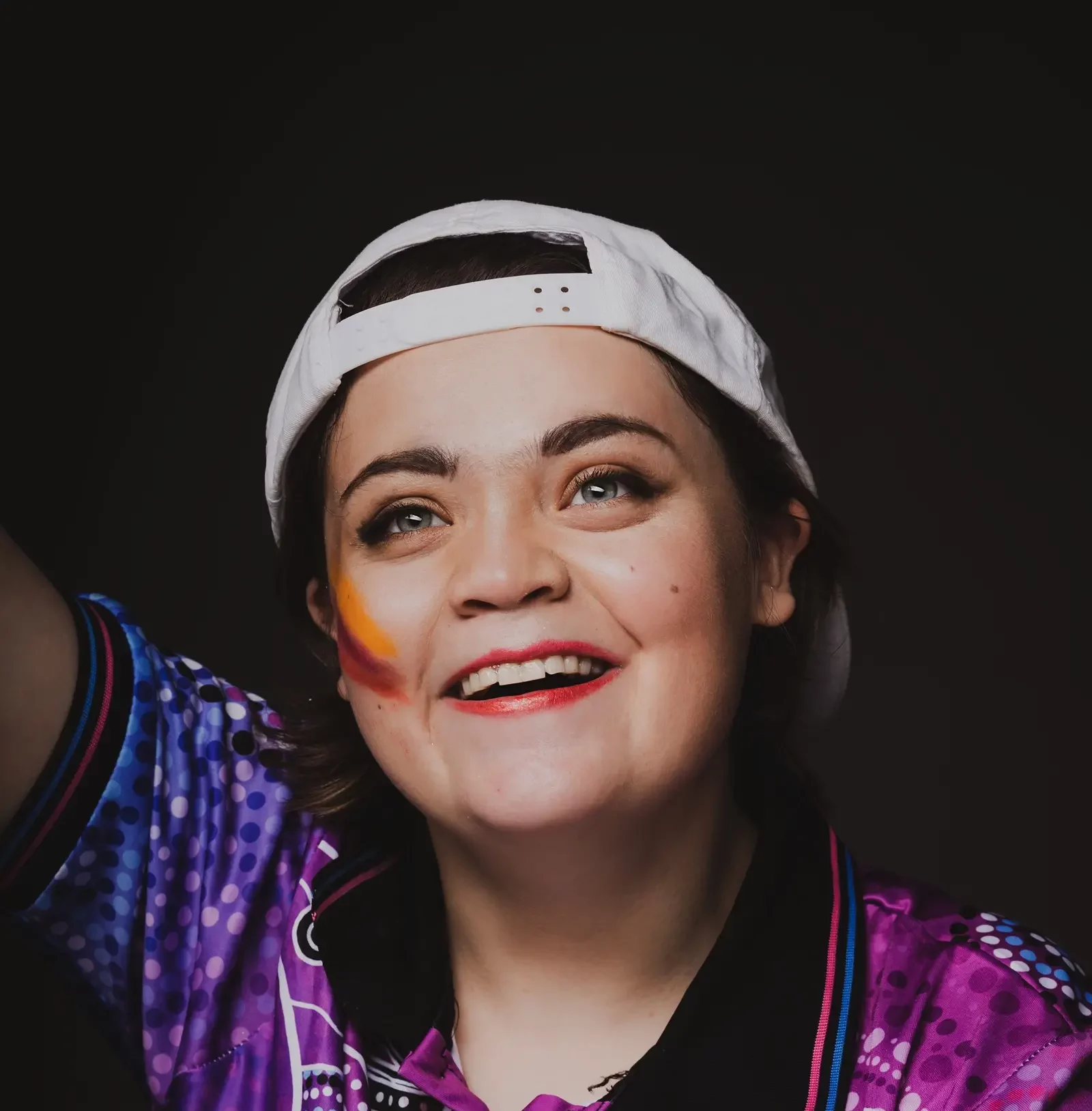 A person with short brown hair wears a backwards white cap and a purple shirt, smiling brightly. They have colourful face paint on one cheek, as if celebrating with friends from Compass Disability Services. The background is dark.