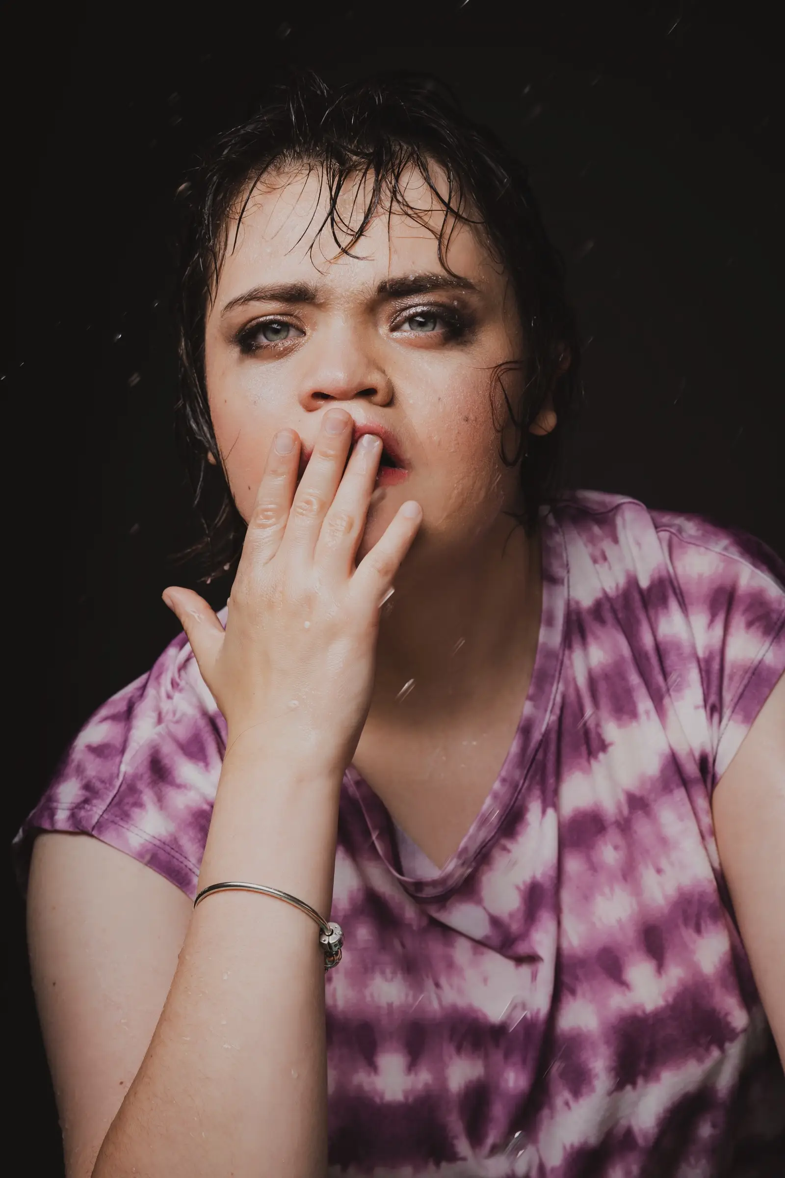 A person with wet hair and dramatic eye make-up wears a purple tie-dye shirt, holding a hand to their mouth against a dark background. Water droplets gleam as if questioning, "What If?"—their gaze a compass pointing towards unseen stories.