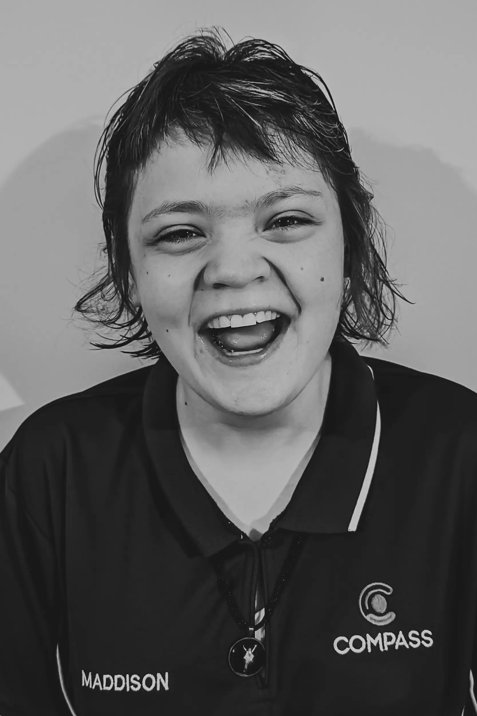 A young person with short, tousled hair smiles widely. They wear a dark collared shirt with the name Maddison and a Compass Disability Services logo on it. The photo is in black and white.