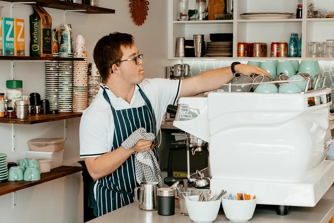 A barista in an apron, demonstrating skills-based training, wipes a cup behind the counter in a coffee shop filled with cups, mugs, and a coffee machine. Shelves with crockery and supplies line the background.