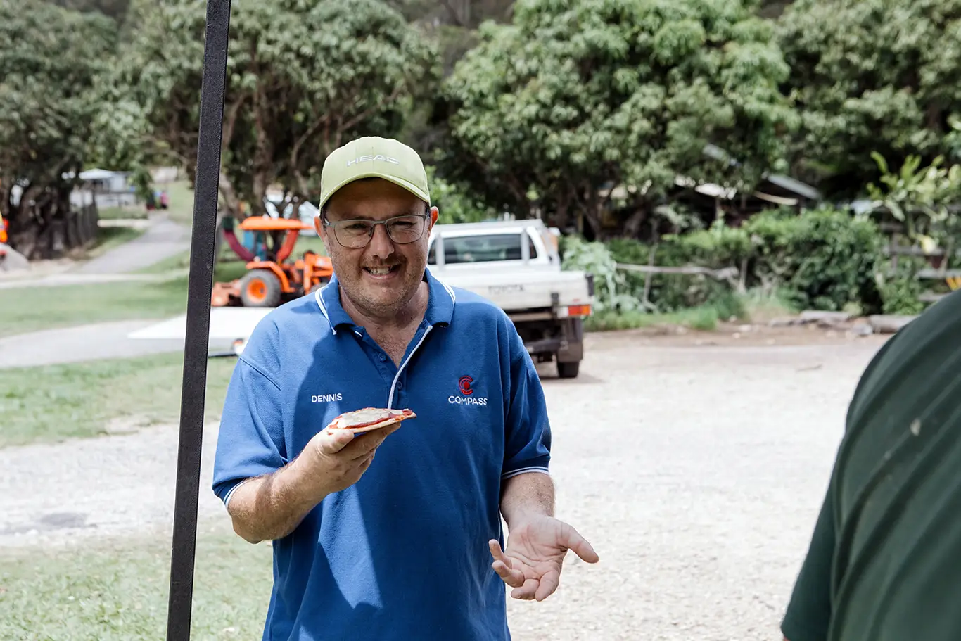 A man in a blue shirt and green cap smiles while holding a slice of pizza outdoors, with trees, a white pickup, and an orange tractor visible—enjoying the moment as if guided by his inner compass.