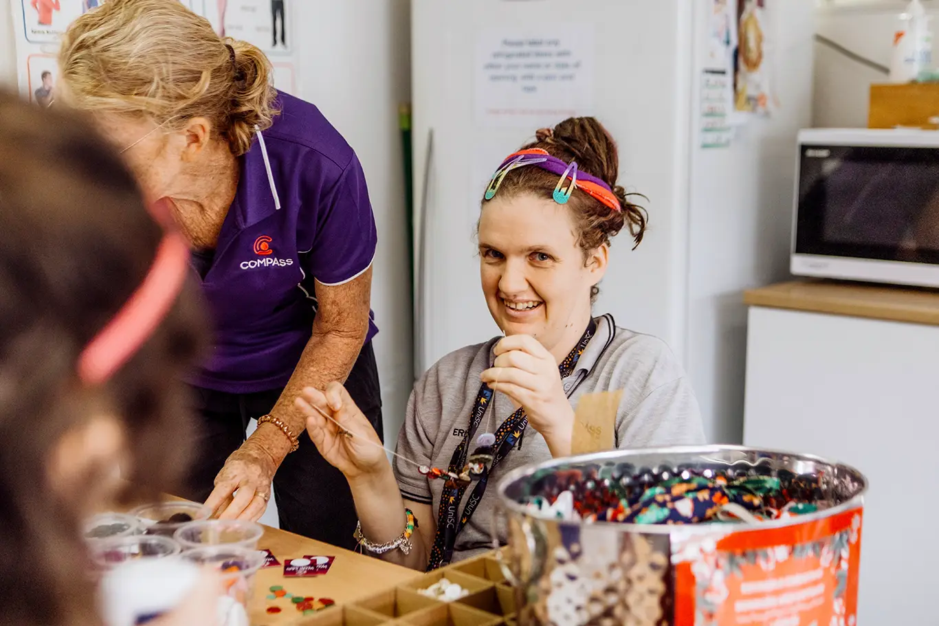 A young woman smiles whilst making crafts with beads at a table, wearing a hairband with colourful hair clips. An older woman from Disability Services stands beside her, helping with the activity. Other supplies and a large bead container are visible.