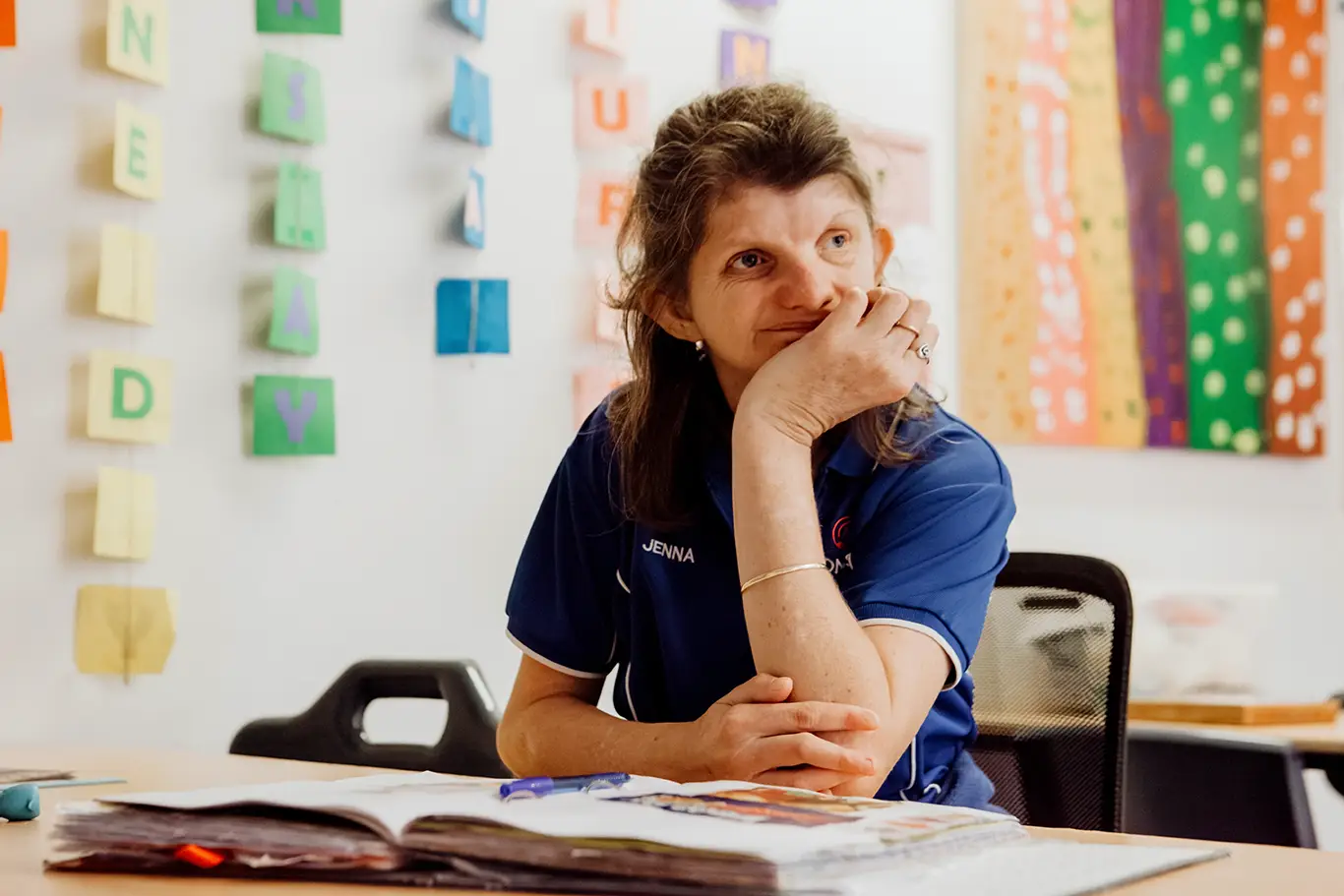 A woman with long hair sits at a desk with an open book, resting her chin on her hand and smiling. Colourful artwork and letters, including the word "Compass," are displayed on the wall behind her.