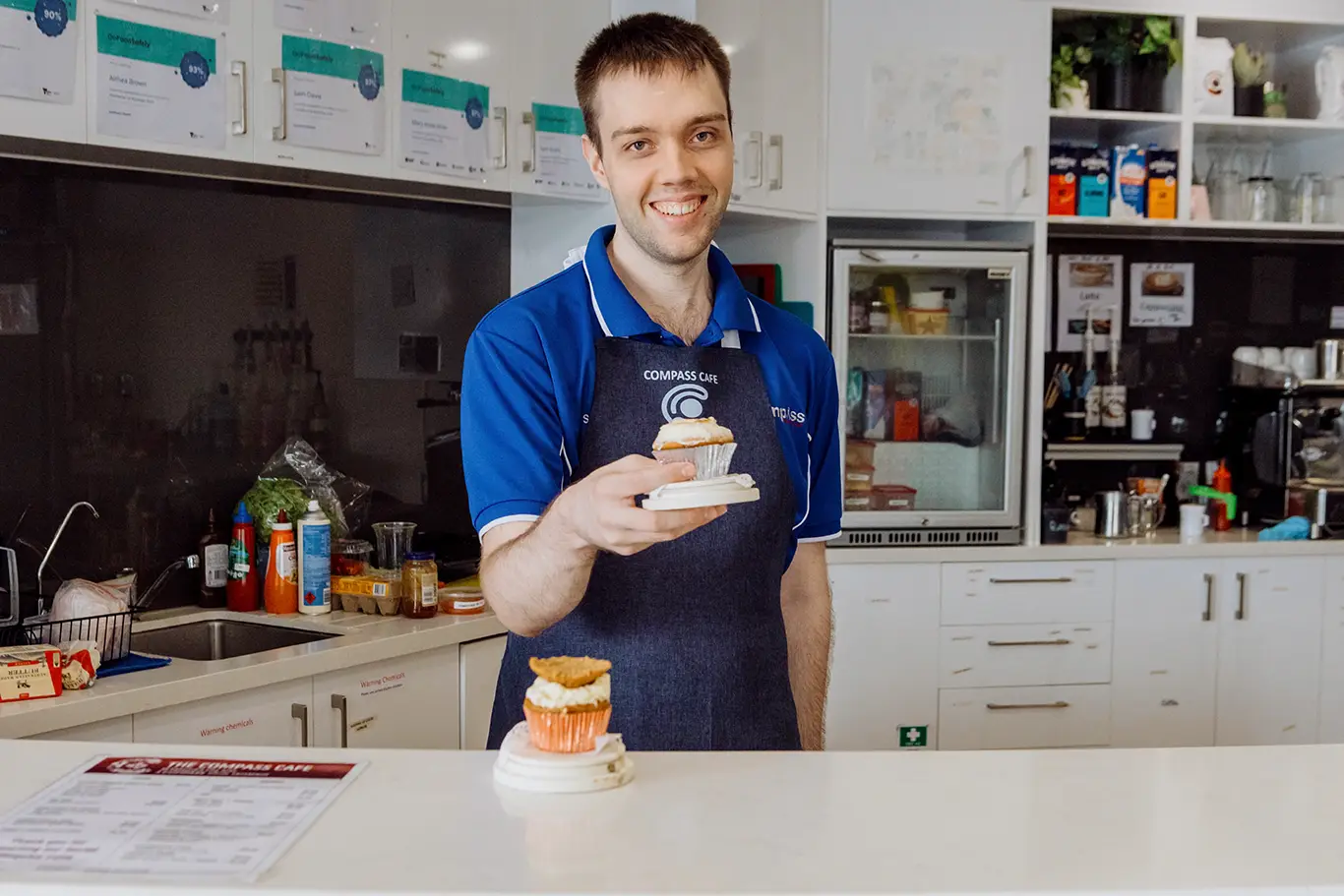 A smiling man in a blue polo shirt and apron stands behind a counter in a café kitchen, holding out a cupcake on a small plate. A "What If" poster about Disability Services hangs on the wall behind him, adding warmth to the welcoming space.