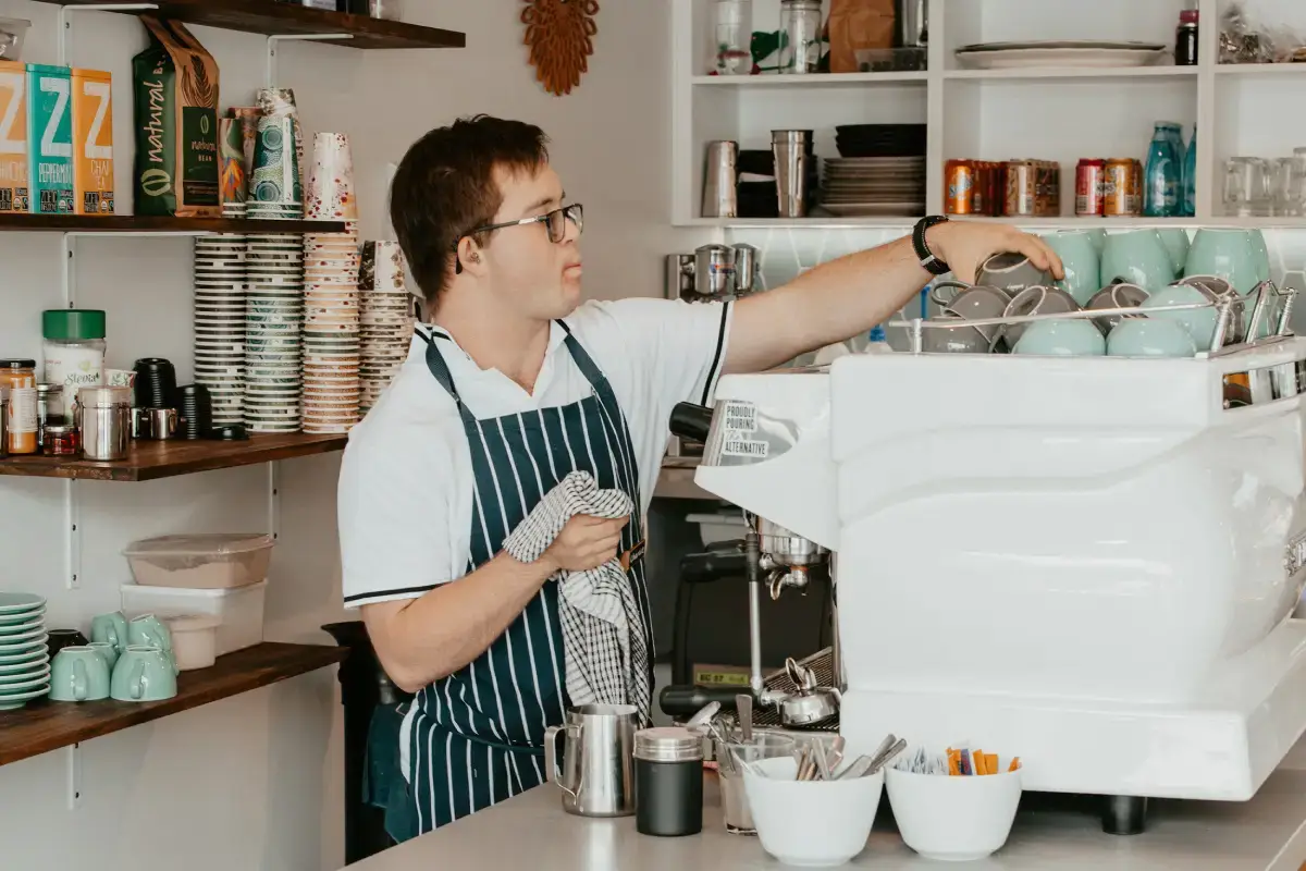 A barista wearing glasses and an apron dries cups behind a coffee machine in a café, surrounded by stacked mugs and various supplies—a quiet moment that feels like a compass guiding the daily rhythm.