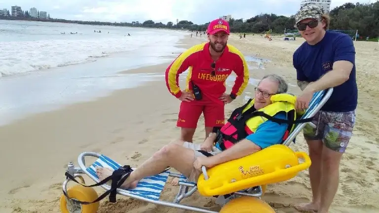 A man in a beach wheelchair provided by Disability Services smiles on the sand, wearing a blue shirt and life jacket. Two men stand beside him—one in a red lifeguard uniform, the other in casual beachwear—with the sea in the background.