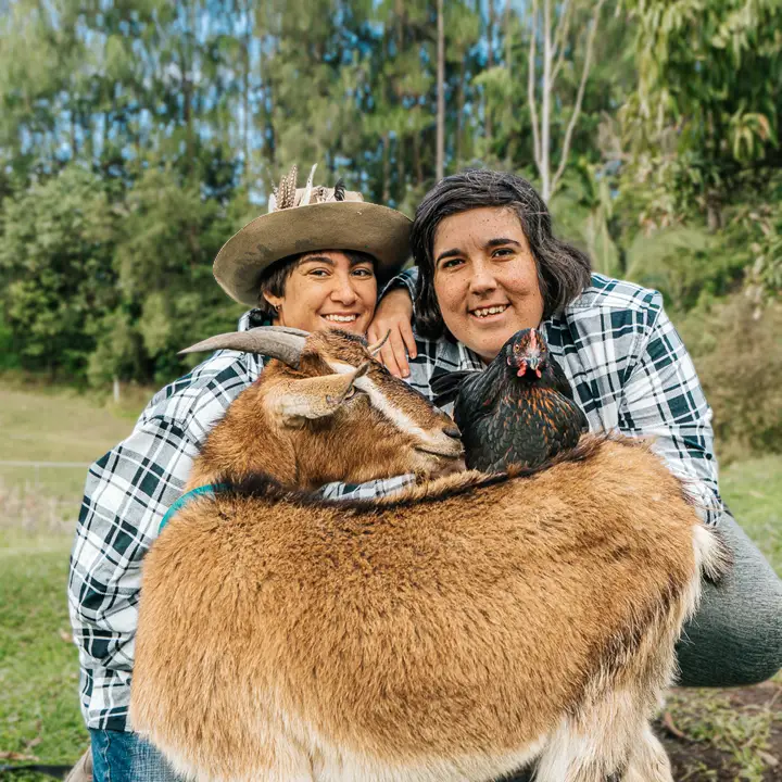 Two people in checked shirts smile while hugging a goat and a chicken outdoors. One person wears a hat with feathers. Surrounded by lush trees, their shared joy sparks the question: what if every day felt this connected to nature?.