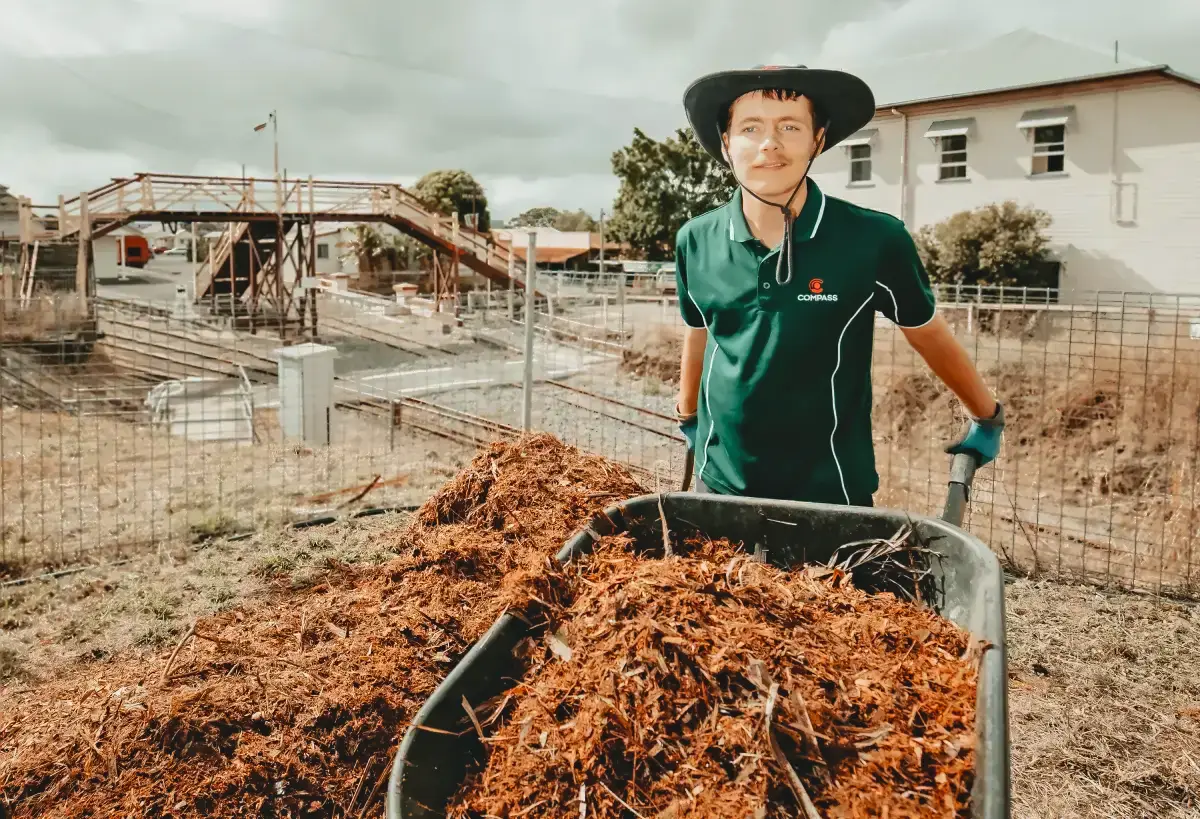 A person wearing a wide-brimmed hat, green shirt, and gloves stands outdoors behind a wheelbarrow filled with mulch, near construction and a building—illustrating how Disability Services can guide individuals as their compass for support.