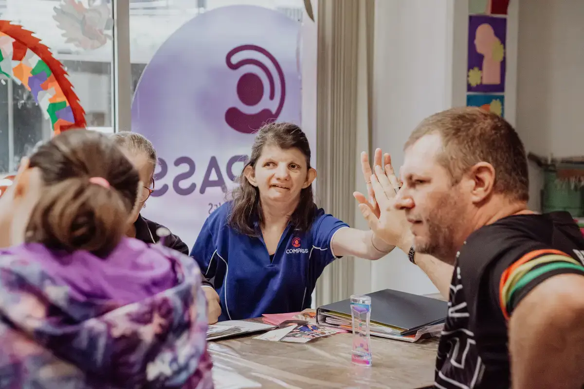 A group of people sit around a table engaging in conversation and activities. One woman in a blue shirt, supported by Disability Services, smiles and raises her hand towards a man in black. Bright decorations and artwork are in the background.