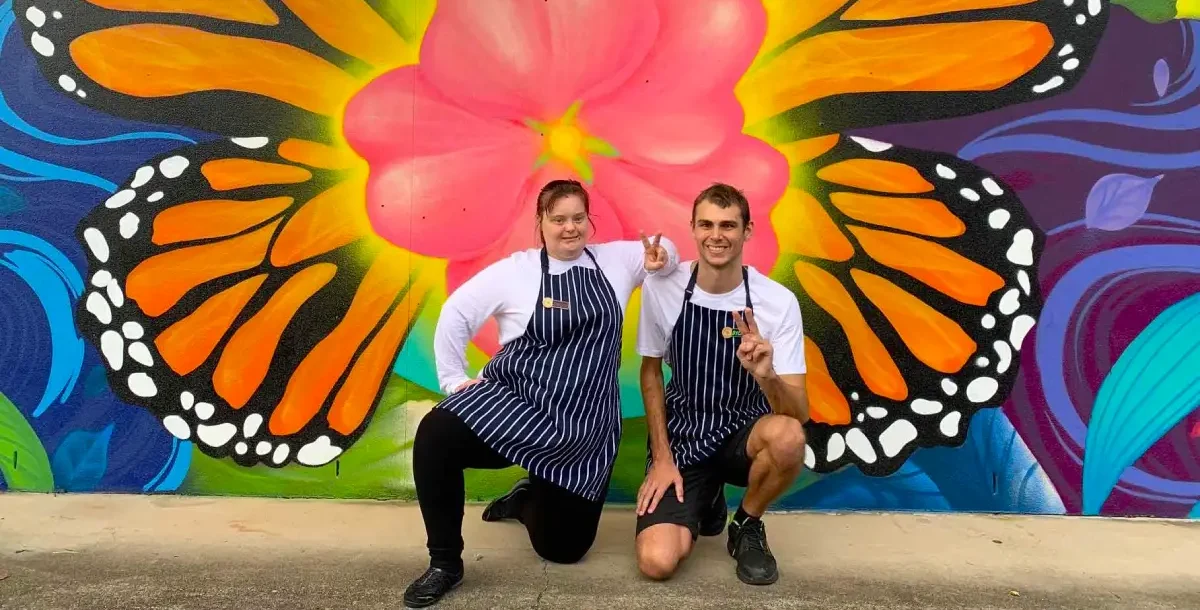 Two people in striped aprons pose in front of a vibrant butterfly mural, their smiles reflecting the positivity fostered by Compass Disability Services. One kneels whilst the other crouches, capturing a joyful "What If" moment together.