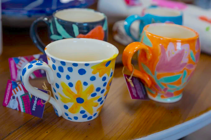 Four colourful ceramic mugs from Wabi Sabi Creations with hand-painted floral and polka dot designs sit on a wooden table. Each mug has a unique pattern and bright colours, with tags attached to the handles.