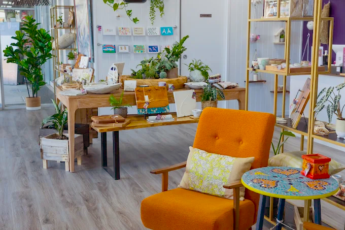 A cosy boutique shop interior by Wabi Sabi Creations, featuring an orange armchair, a colourful round table, wooden display tables with plants, books, and crafts, plus shelves of art and cards against white walls and wood flooring.
