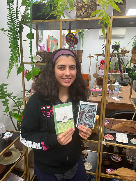 A woman with dark hair and a pink hairband smiles whilst holding two handmade cards from Wabi Sabi Creations in a craft shop filled with plants, shelves, and various art pieces.
