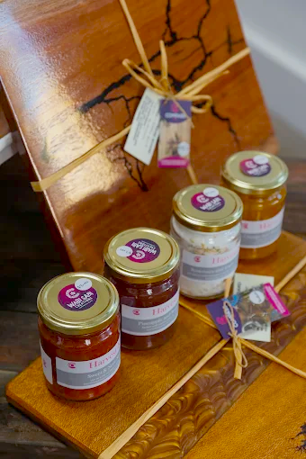 Four jars of homemade preserves from Wabi Sabi Creations with gold lids are arranged on two carved wooden boards. Decorated and tied with string, each board holds jars featuring charming white and red labels with attached tags.