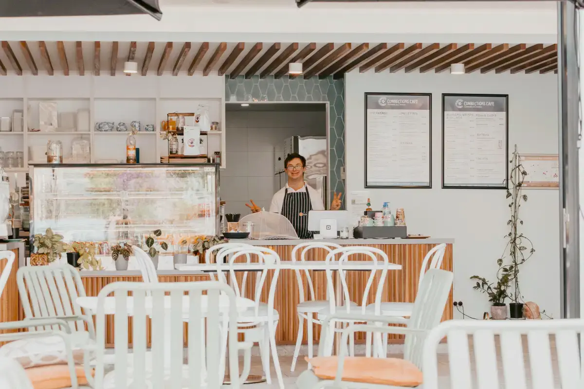 A person wearing an apron stands behind the counter of Connections Café Maroochydore, smiling and holding up two peace signs. White chairs and tables fill the foreground, and menus are displayed on the back wall of this bright, modern café.