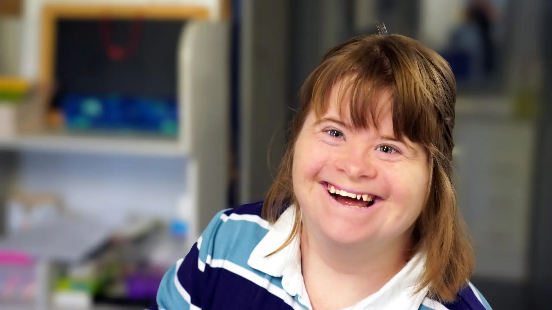 A young woman with Down's syndrome smiles warmly at the camera. She has straight, light brown hair with a fringe and is wearing a blue and white striped shirt. The background is softly blurred, showing an indoor setting.