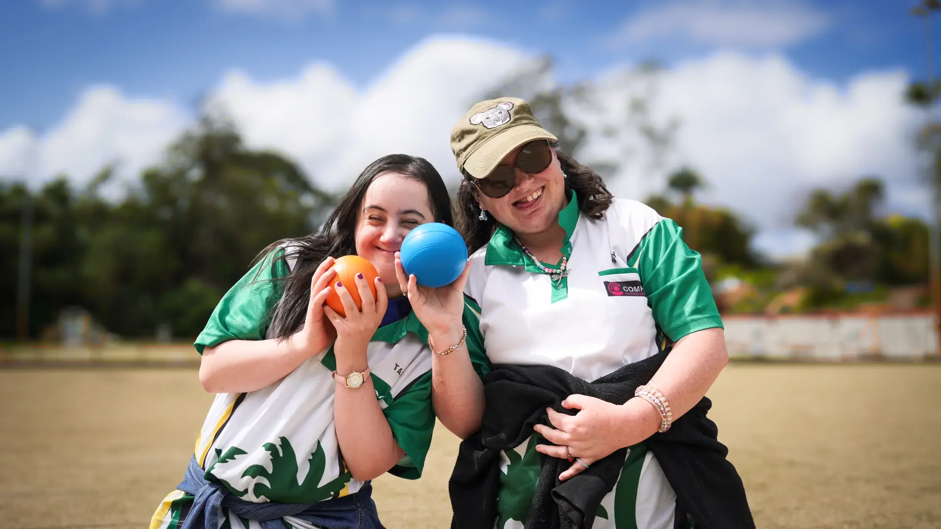 Two women smiling outdoors, wearing green and white shirts, each holding a colourful ball. One woman wears a cap and sunglasses; both appear happy and are posing together on a sunny day.