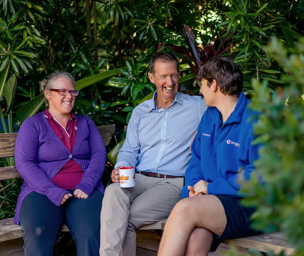 Three people sit on a wooden bench outdoors, surrounded by greenery, smiling and chatting. One holds a coffee mug as they enjoy a friendly conversation—capturing the spirit of our about us moments.
