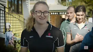 A smiling woman with glasses and a ponytail, wearing a black COMPASS polo shirt, stands outdoors in front of three other people in uniforms—highlighting opportunities for careers and volunteering near a building with steps and greenery.