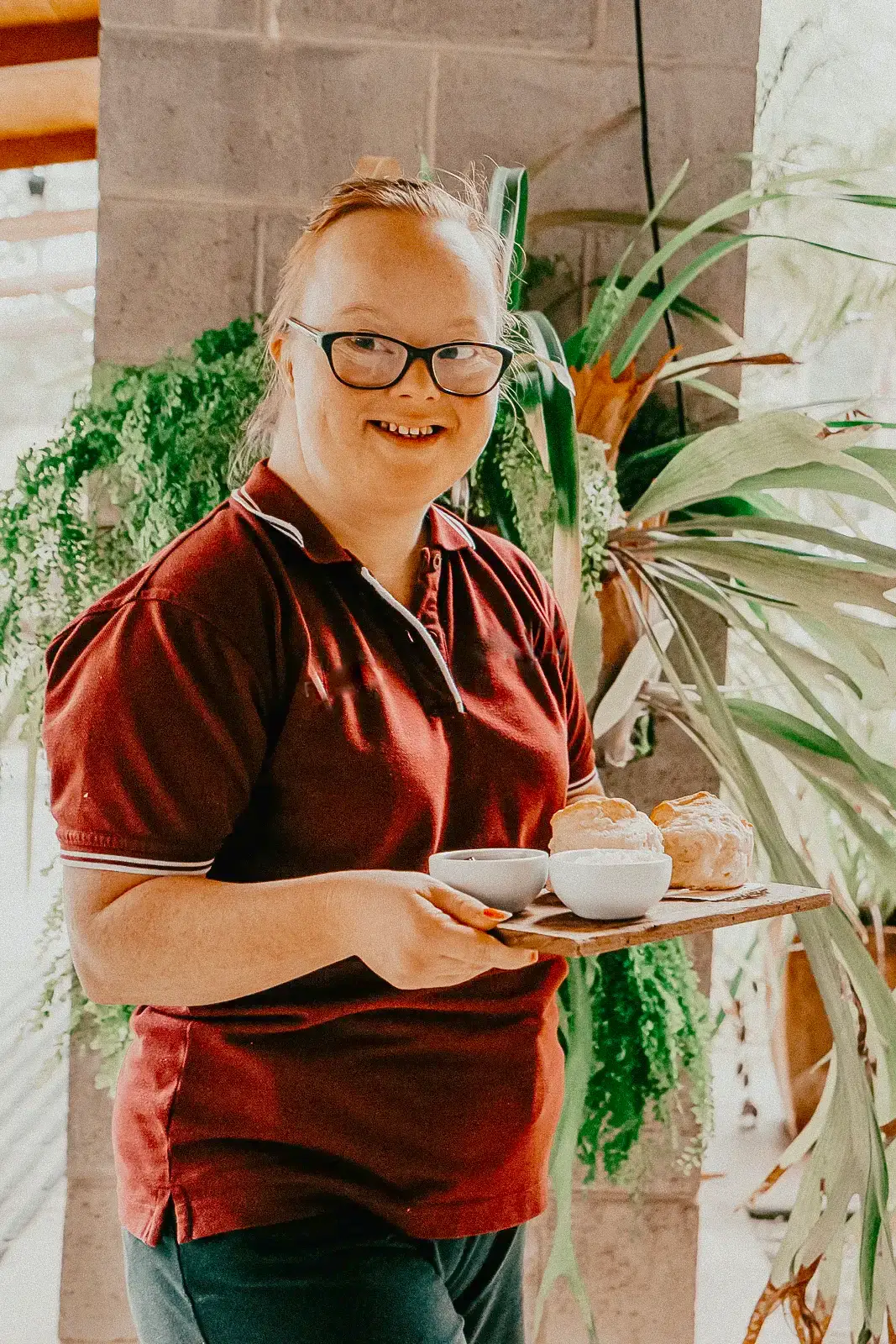 A smiling person wearing glasses and a maroon shirt holds a tray with two mugs and pastries, standing in front of lush green plants indoors at the Garden Café.