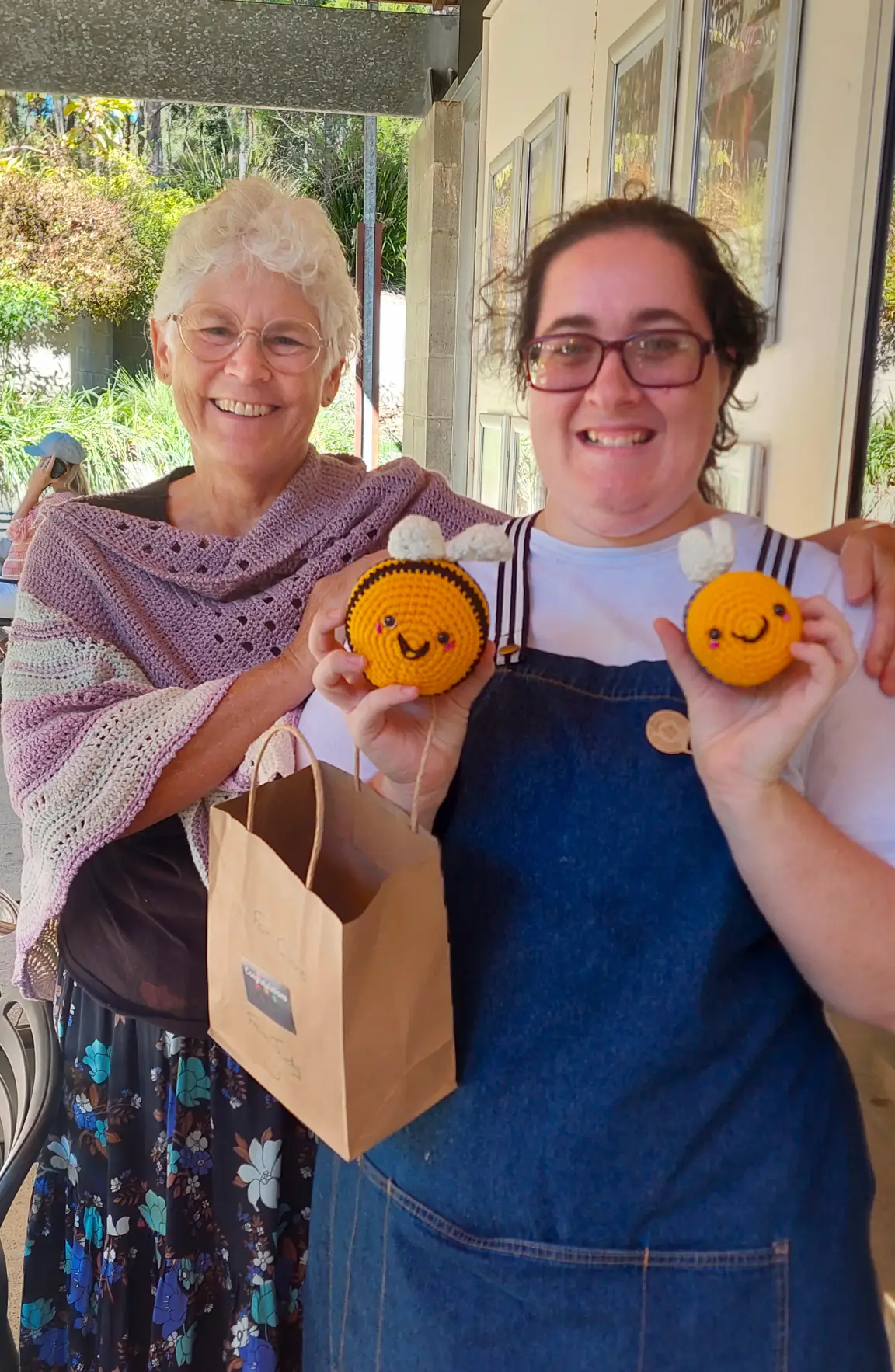 Two smiling women pose together at a Garden Café; one holds a paper bag, while the other shows off two crocheted bees with happy faces. Sunlight brightens the cosy, outdoor café setting.