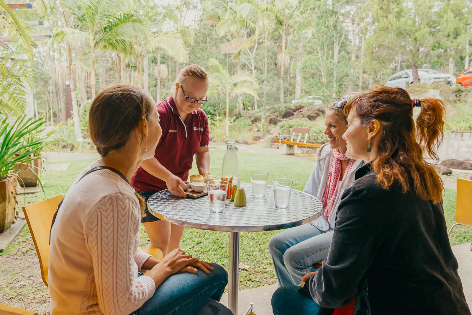 A waiter brings food to three women sitting at a Garden Café table surrounded by greenery. The women are smiling and chatting, with water glasses and condiments on the table.