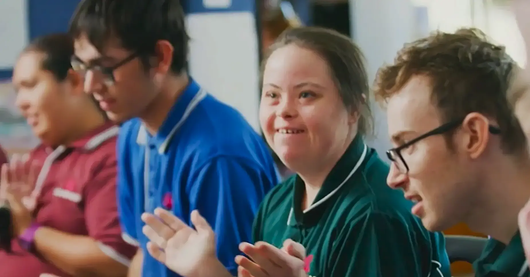 A group of young people with disabilities, wearing coloured polo shirts, sit together and clap, smiling and enjoying themselves in a bright indoor setting—promoting inclusion in your community.